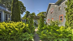A pathway flanked by lush greenery and a wooden pergola, leading to a house with a grey facade and red-framed windows. - Walk to Dtwn Shops & Eats: Sturgeon Bay Retreat (Sturgeon Bay)
