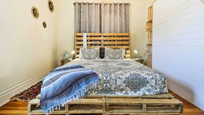 A bedroom with a wooden pallet bed, patterned bedding, a hanging light fixture, and a bedside table with a lamp. - Walk to Dtwn Shops & Eats: Sturgeon Bay Retreat (Sturgeon Bay)