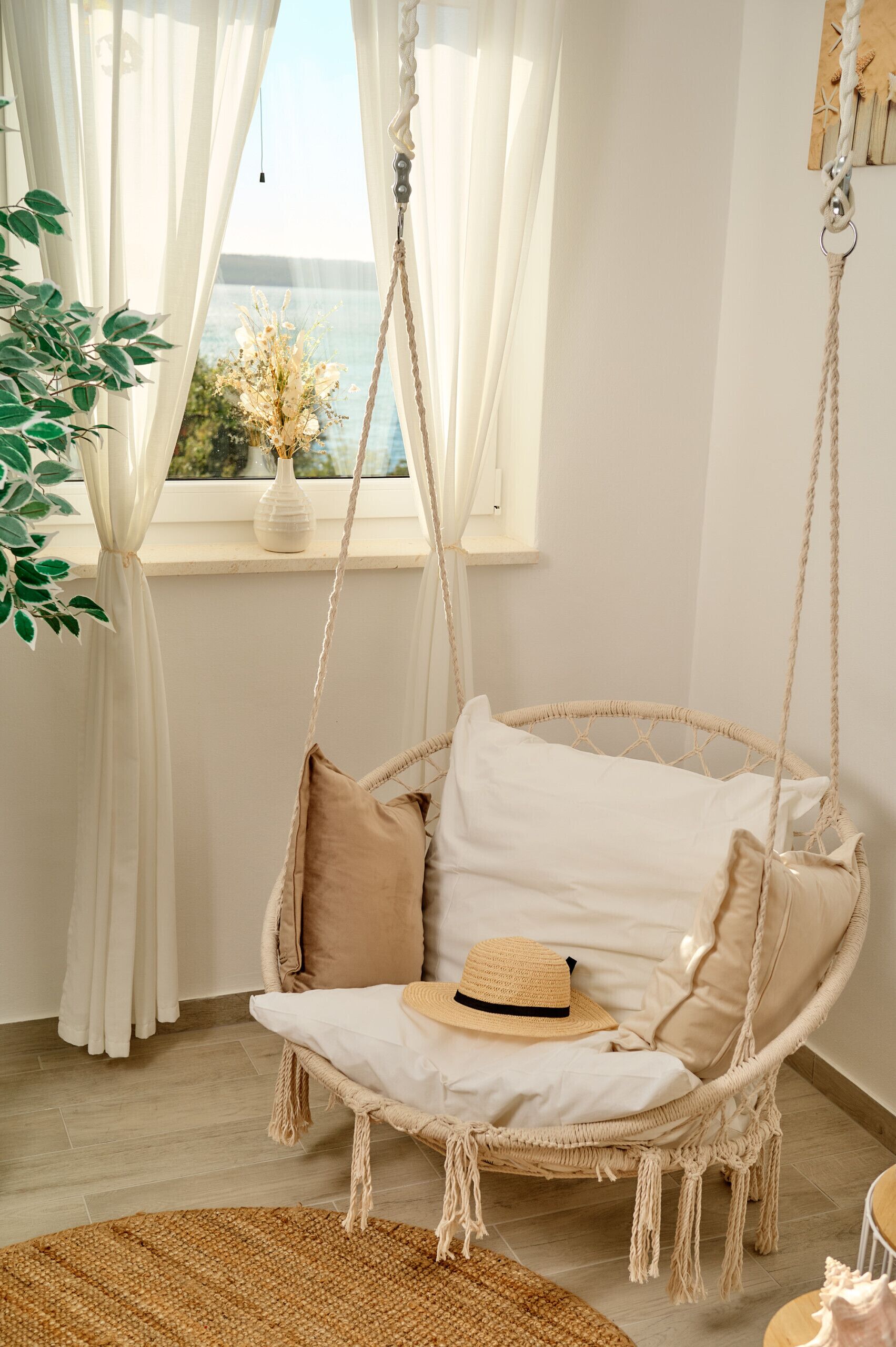 Una silla mecedora de mimbre con cojines, un tapete tejido y vistas al exterior a través de una ventana con cortinas blancas.