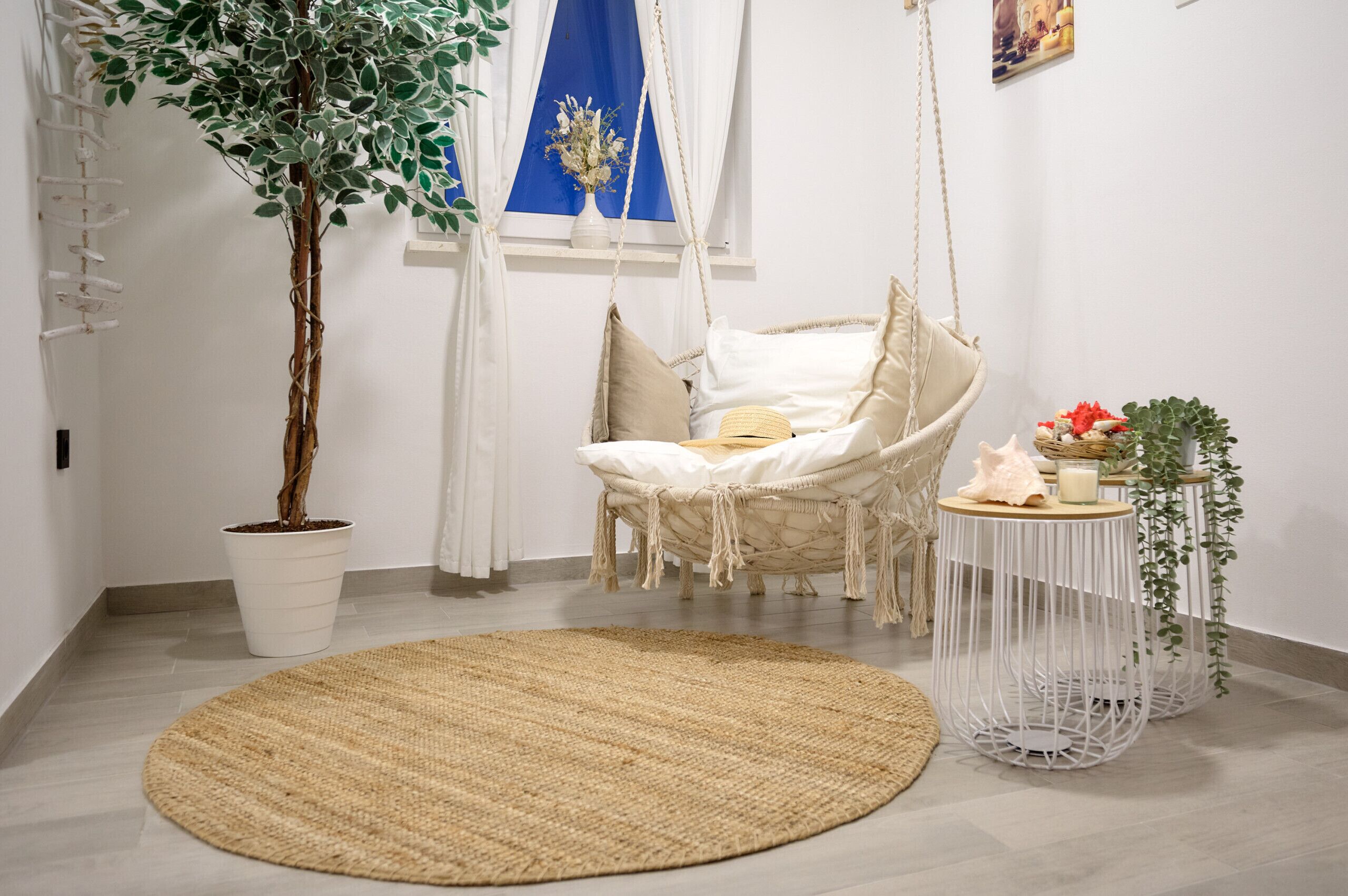 Ein Zimmer mit Hängesessel, Topfpflanze, rundem Teppich und kleinem Tisch mit Dekoration.