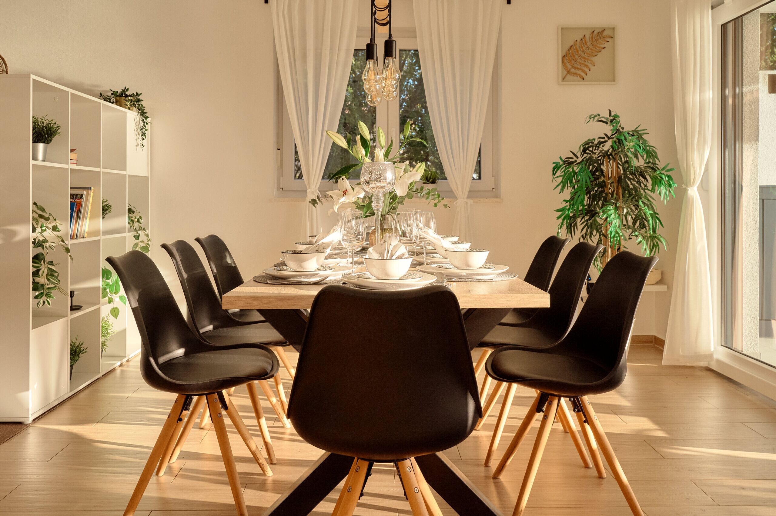 Een eetgedeelte met een houten tafel voor zes personen, omringd door zwarte stoelen met houten poten. Links een witte wandplank met planten en rechts een groot raam met witte gordijnen.