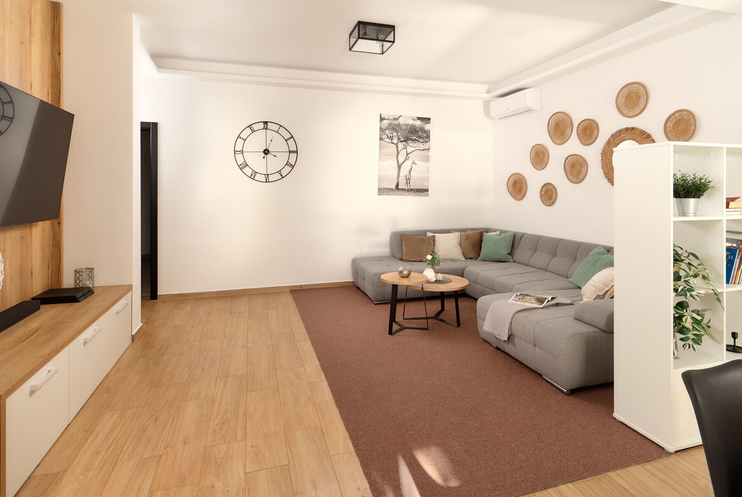 Ein modernes Wohnzimmer mit grauem Sofa, einem Couchtisch, einem Flachbildfernseher, einer Wanduhr und einem Bücherregal.