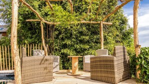 A wooden pergola structure with wicker furniture, including two sofas and a table, set against a backdrop of lush greenery.
