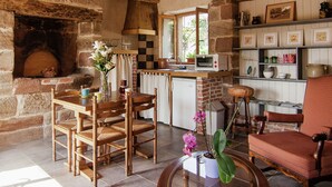 A rustic kitchen with a brick oven, wooden furniture, and a window overlooking greenery.