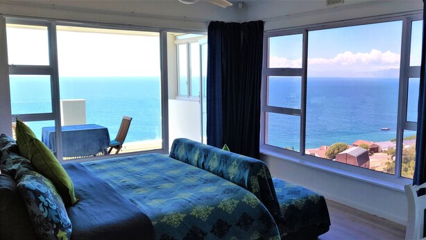 A room with a bed, a chair, and a view of the sea through large windows.