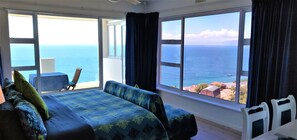 A room with a bed, a chair, and a view of the sea through large windows.