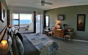 A bedroom with a bed, a desk, a chair, a dresser, and a view of the ocean through the balcony doors.