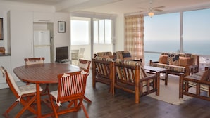 A spacious living area with a wooden dining table and chairs, a sofa set, and a large window offering a view of the ocean.