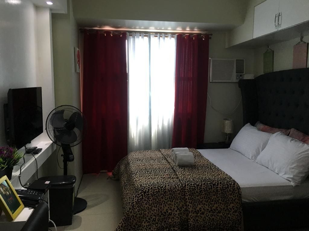 Une chambre d’hôtel avec un lit, un bureau avec un ordinateur, un ventilateur et une fenêtre avec des rideaux rouges.