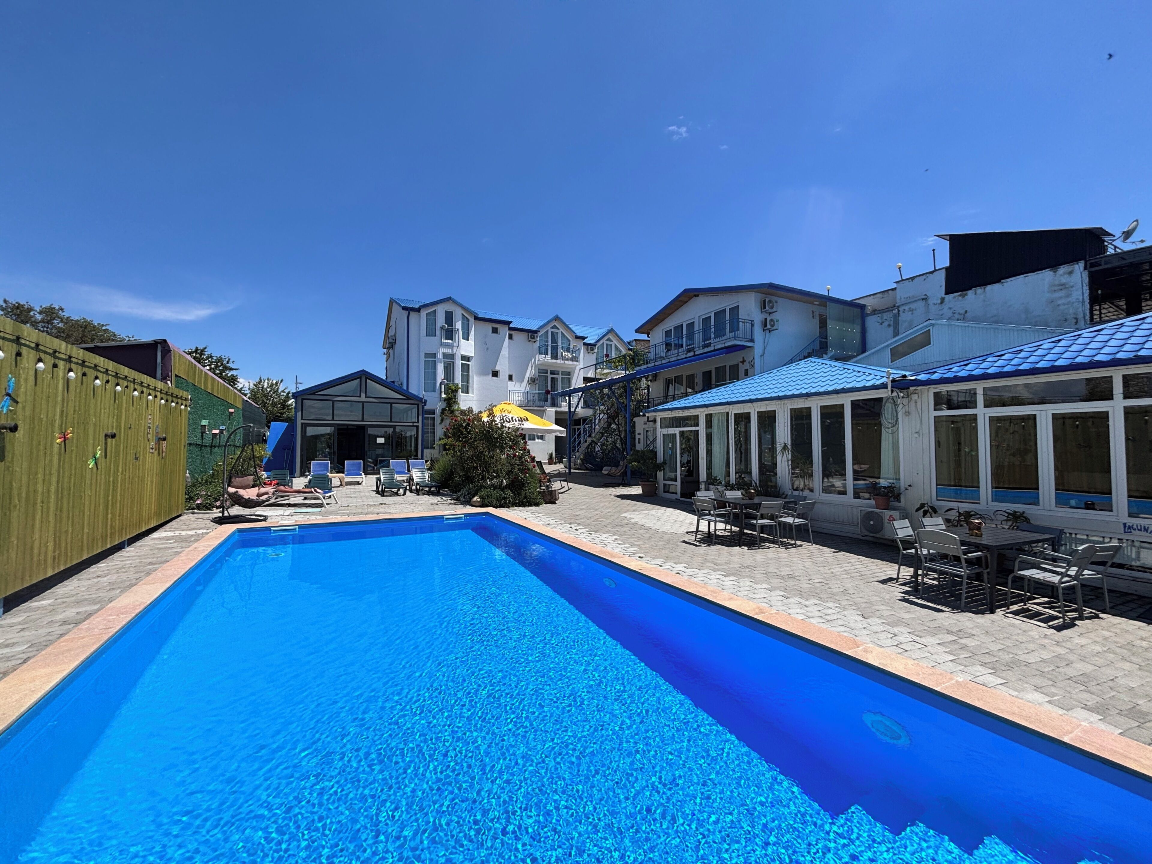 En swimmingpool med solstolar, en möblerad uteplats under tak och en byggnad med ett blått tak.