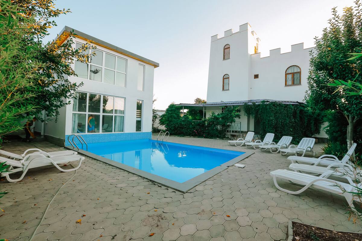 Una piscina con sillones reclinables, un edificio moderno y una estructura que recuerda a un castillo.