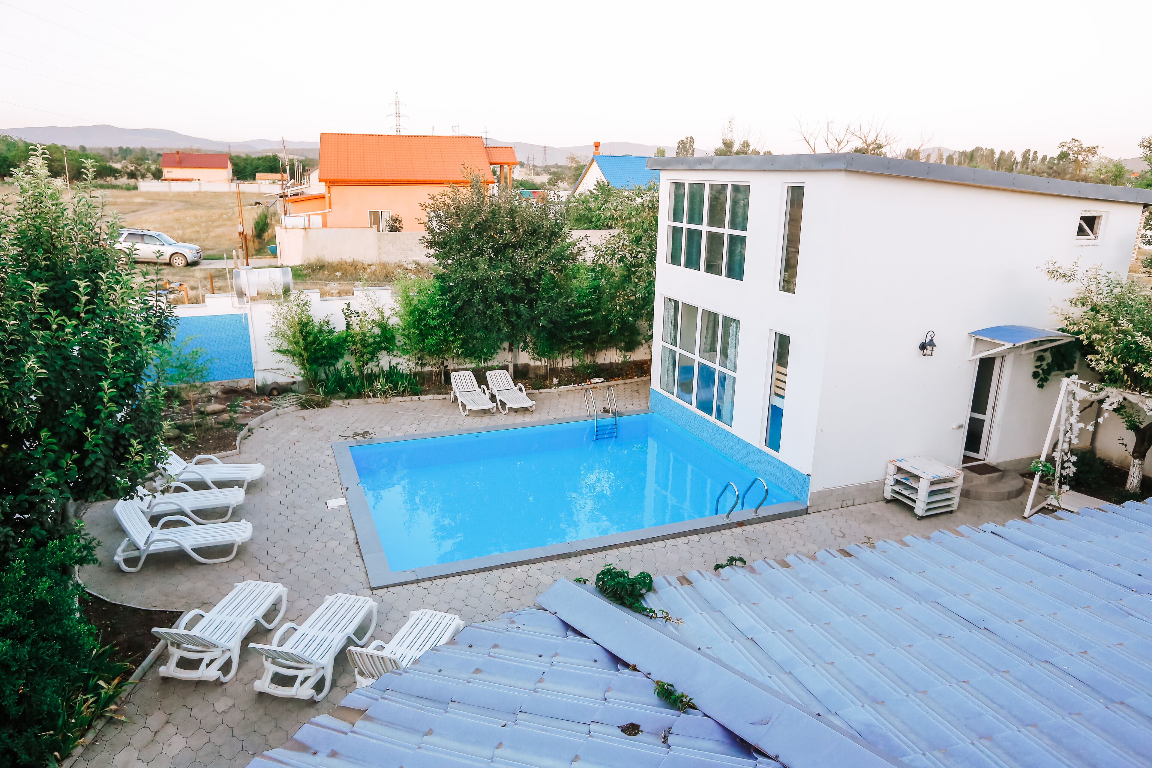 A swimming pool with lounge chairs, a white building with large windows, and a red-roofed house in the background.