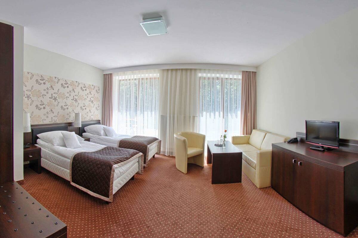 Ein Hotelzimmer mit zwei Betten, einem Schreibtisch, einem Sessel, einem Fernseher und einem großen Fenster mit Vorhängen.