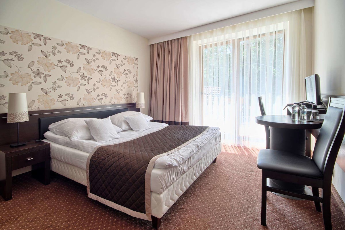 Ein Hotelzimmer mit Bett, Schreibtisch, Stuhl, Lampe und einem fenster mit Vorhängen.