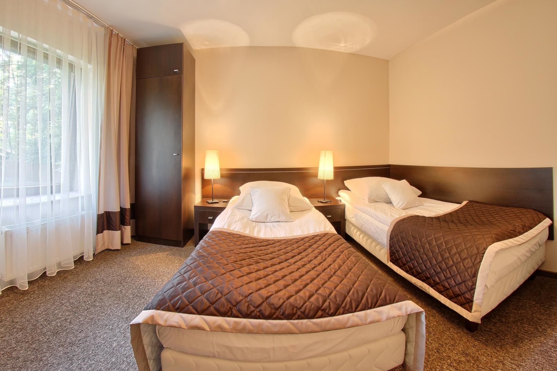 A hotel room with two beds, a window with sheer curtains, and a nightstand with a lamp.
