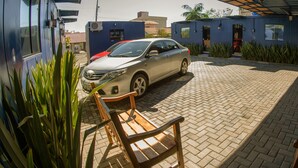 A car parked under a covered area with a brick floor, a wooden bench, and a building with a blue exterior.