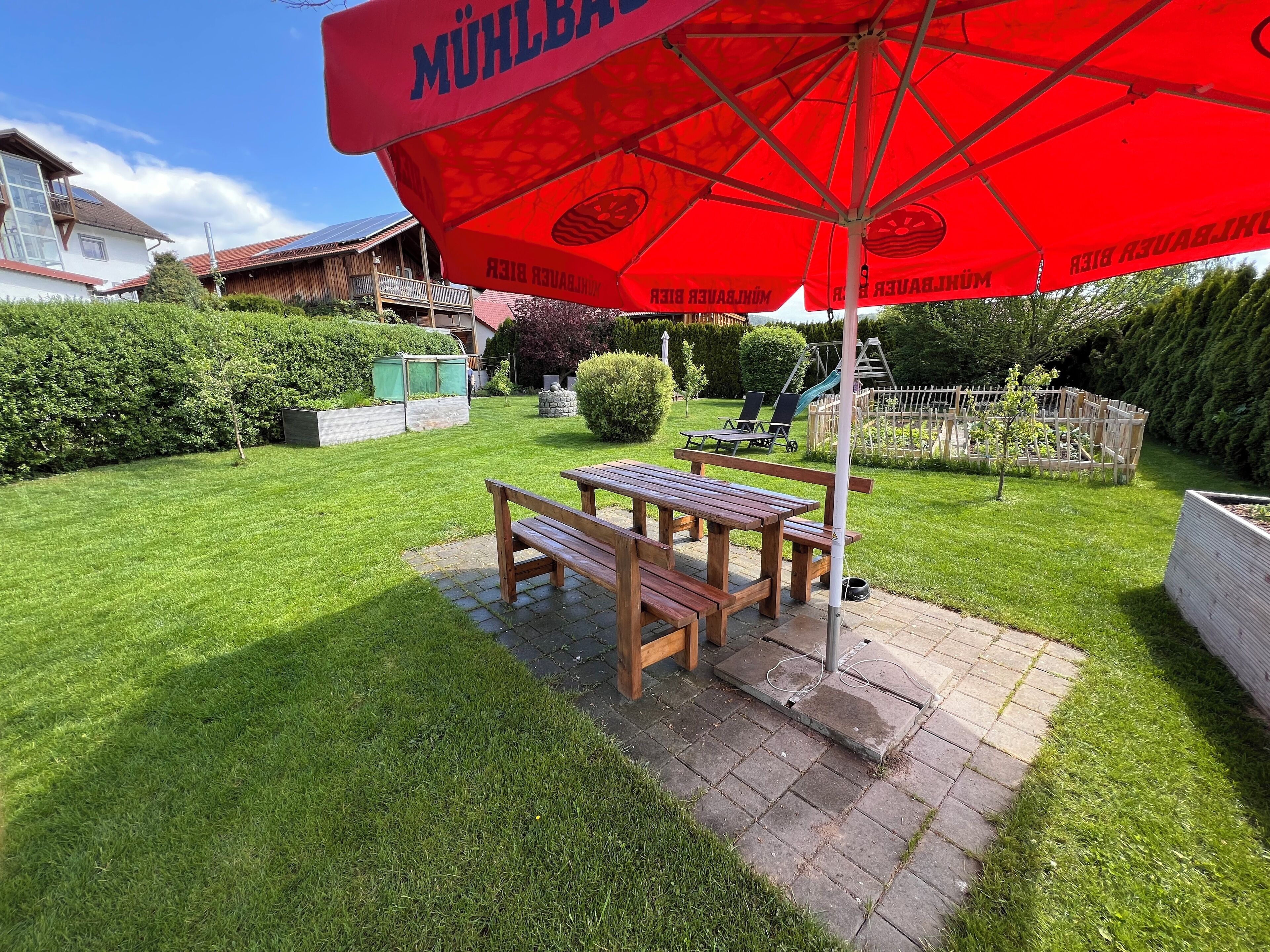 Un patio con una mesa y bancos de madera, un paraguas rojo y un área de juegos al fondo.