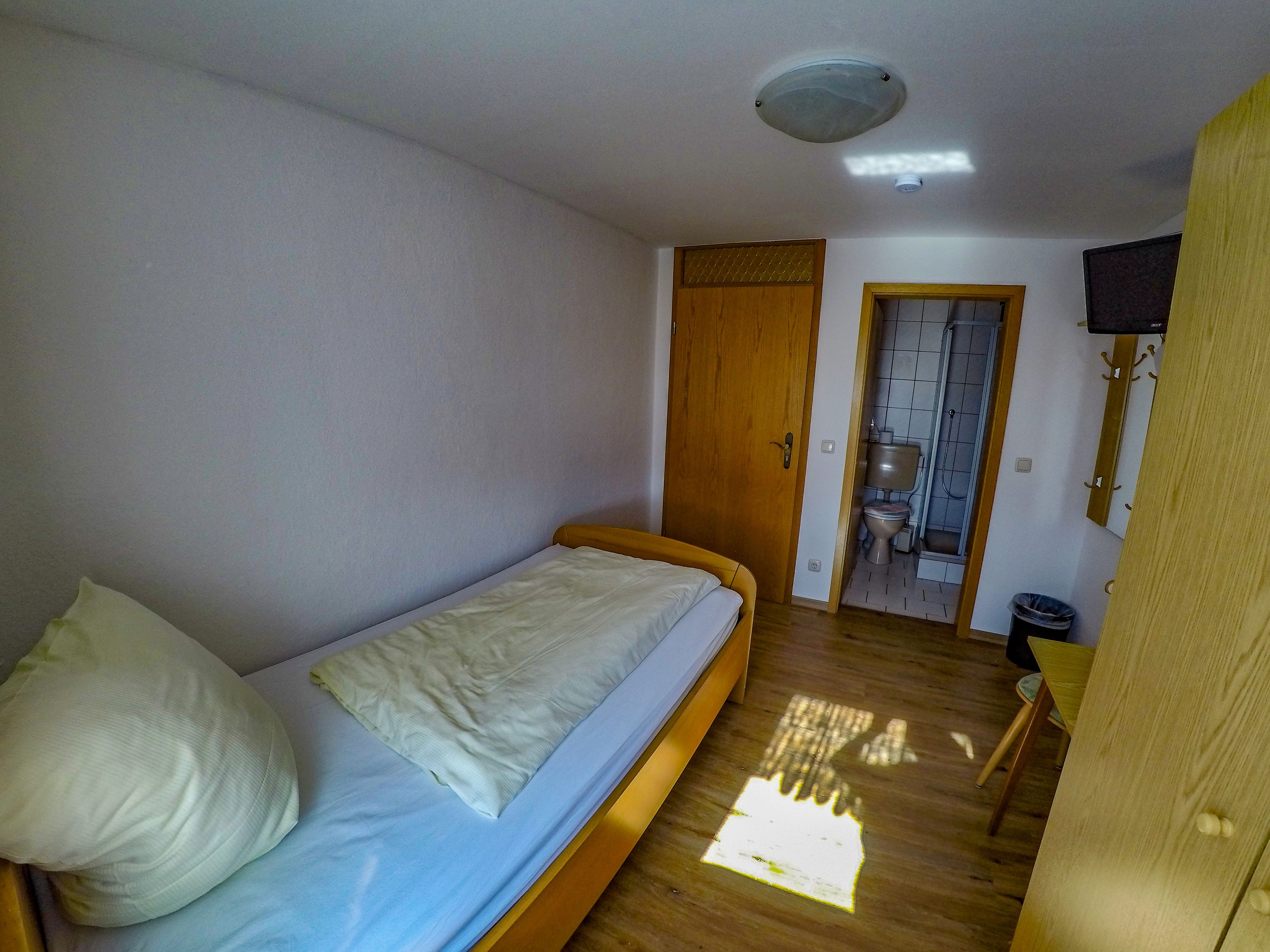 Una habitación pequeña con cama individual, piso de madera, puerta de madera y una mesa pequeña con una silla.