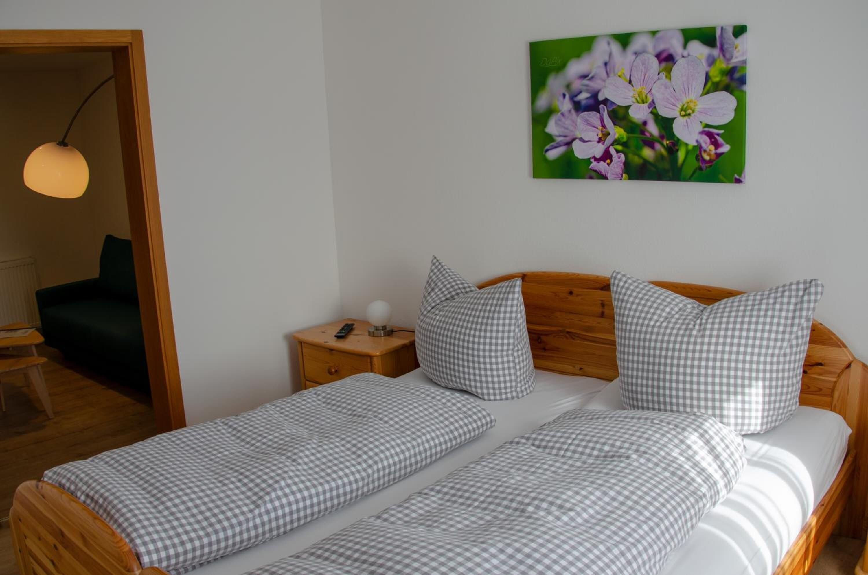 A bedroom with two single beds, a wooden headboard, a nightstand with a lamp, and a wall-mounted picture of flowers.