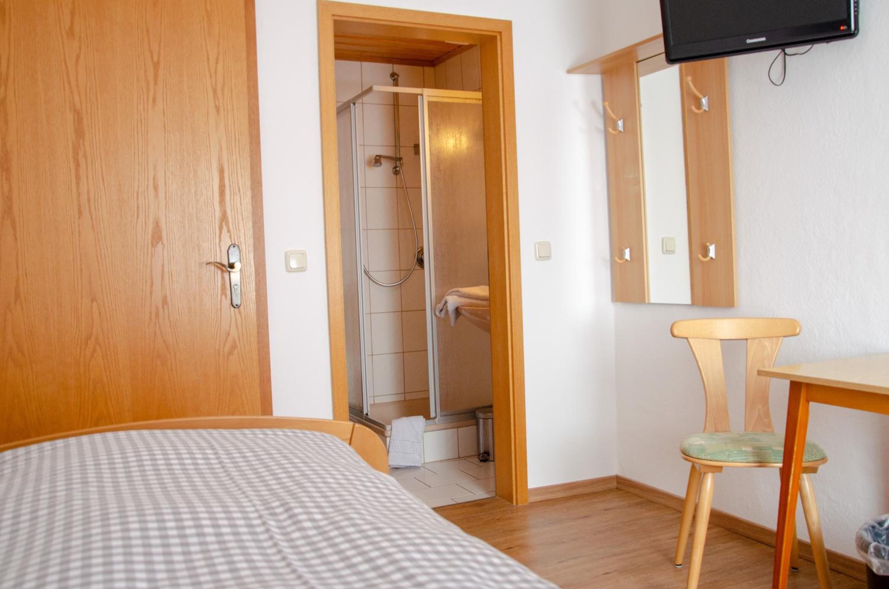 A bedroom with a bed, a wooden door, a shower area, a wooden chair, and a television on the wall.