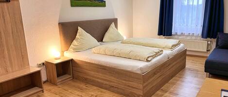 Un dormitorio con una cama grande, una mesita de noche con lámpara, un cabecero de madera, un cuadro en la pared y una ventana con cortinas.