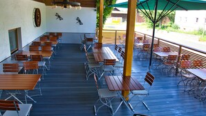 An outdoor dining area with multiple wooden tables and metal chairs, a green umbrella, and a wooden ceiling. - Gasthof Altmann (Eschlkam)