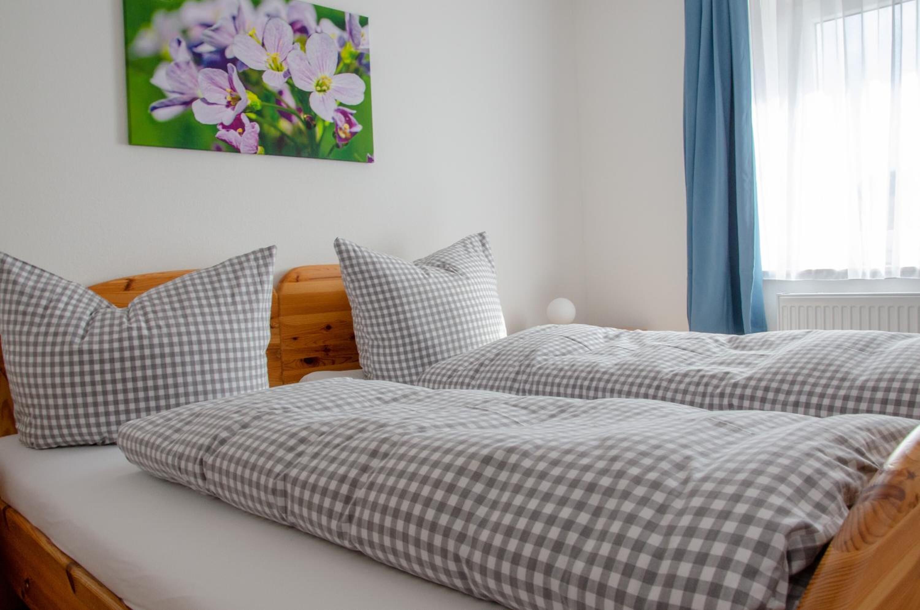 Quarto com cabeceira de madeira, duas camas com roupa de cama xadrez, um quadro de flores na parede e uma janela com cortinas azuis.