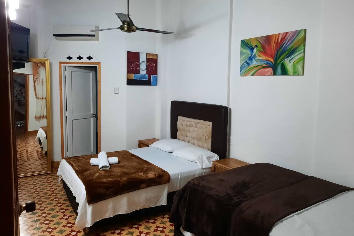 Habitación de hotel con cama, ventilador de techo, mesita de noche pequeña y aire acondicionado de pared.