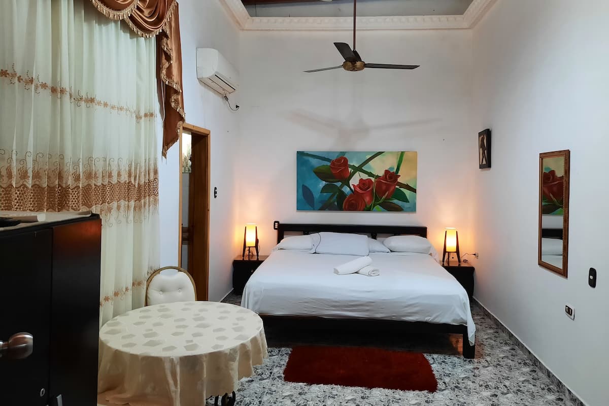 Un dormitorio con cama, dos lámparas de noche, un ventilador de techo, un cuadro de rosas y una mesa con una silla.