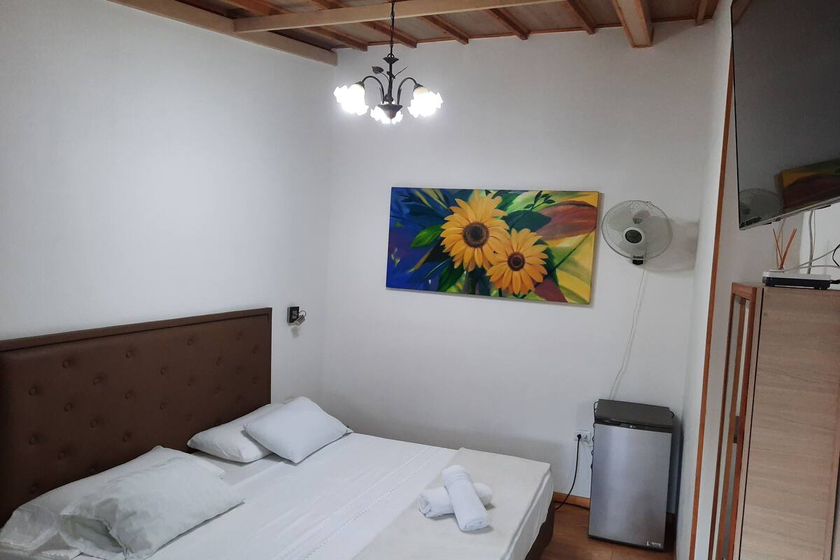 Un dormitorio con una cama, un ventilador de pared, un cuadro de girasoles y un televisor sobre un mueble de madera.