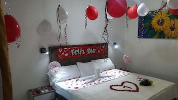 Una cama con pétalos de rosa y una decoración con forma de corazón, una mesita de noche con un teléfono, y una pared con un cartel que dice 'Feliz Dia'.