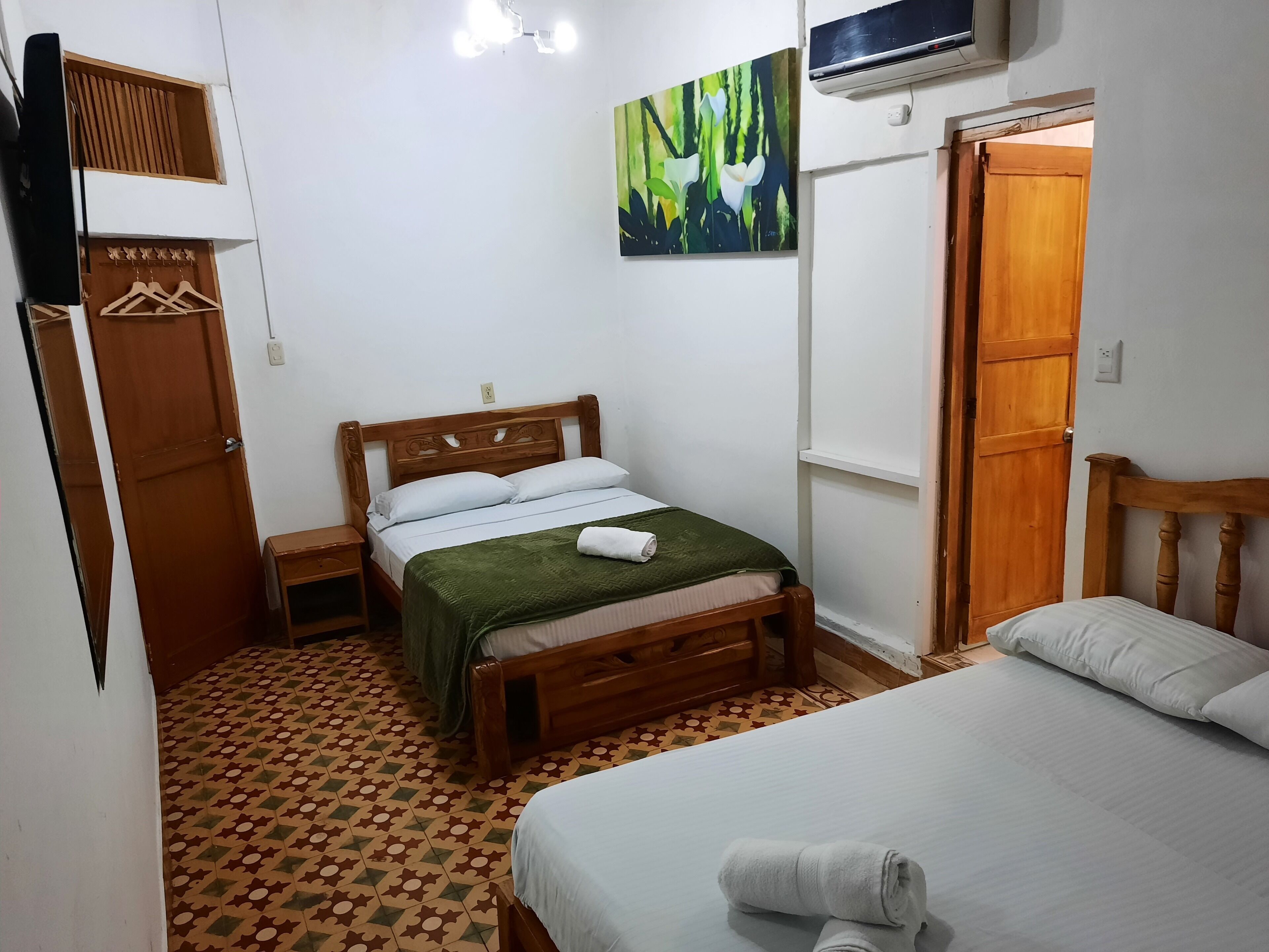 A bedroom with a wooden bed, a green blanket, and a white pillow. There is a painting on the wall and an air conditioning unit above the bed.