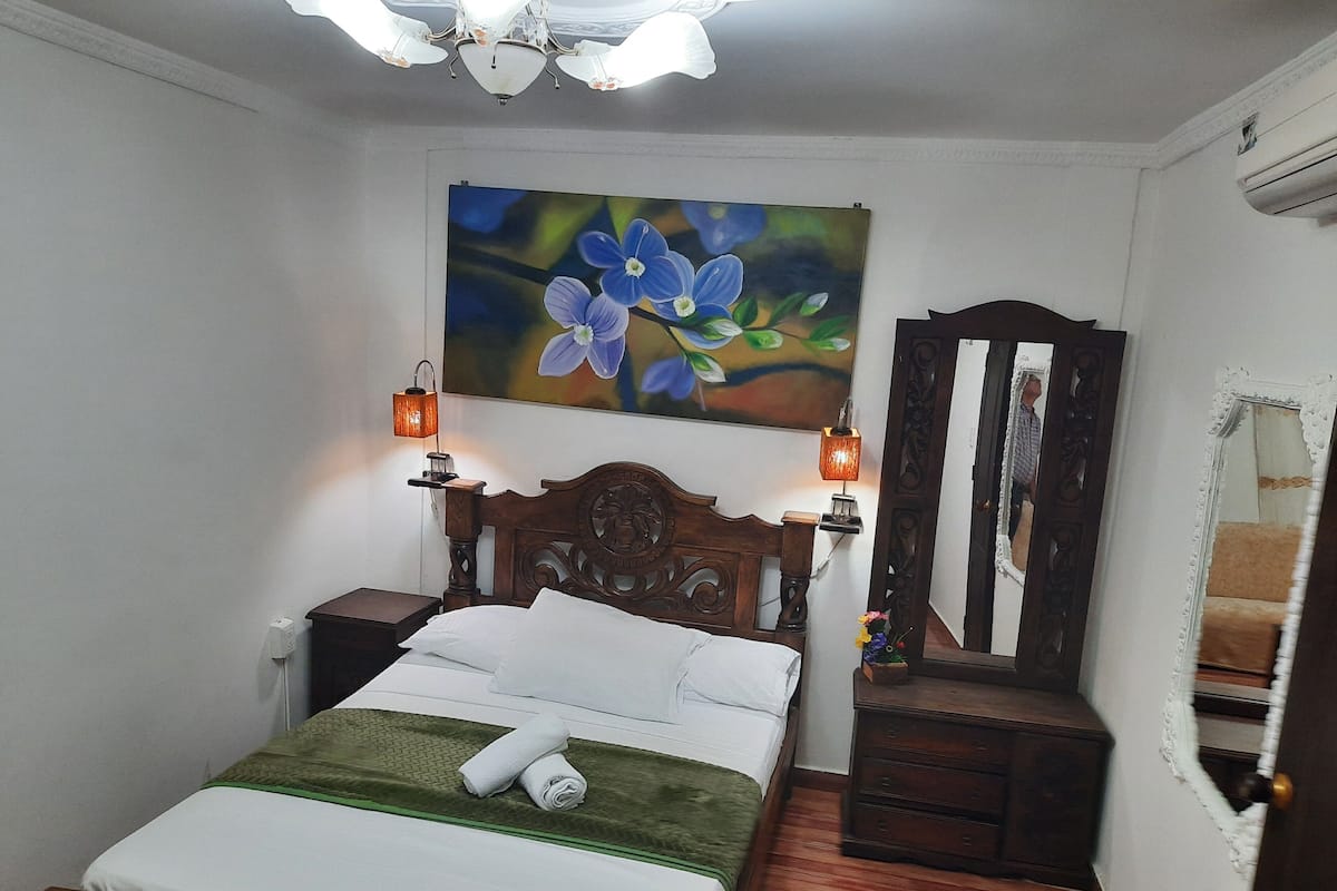 Una cama bien hecha con una colcha verde, dos lámparas de noche, un cabecero de madera, un cuadro de flores, un aparador y un espejo.