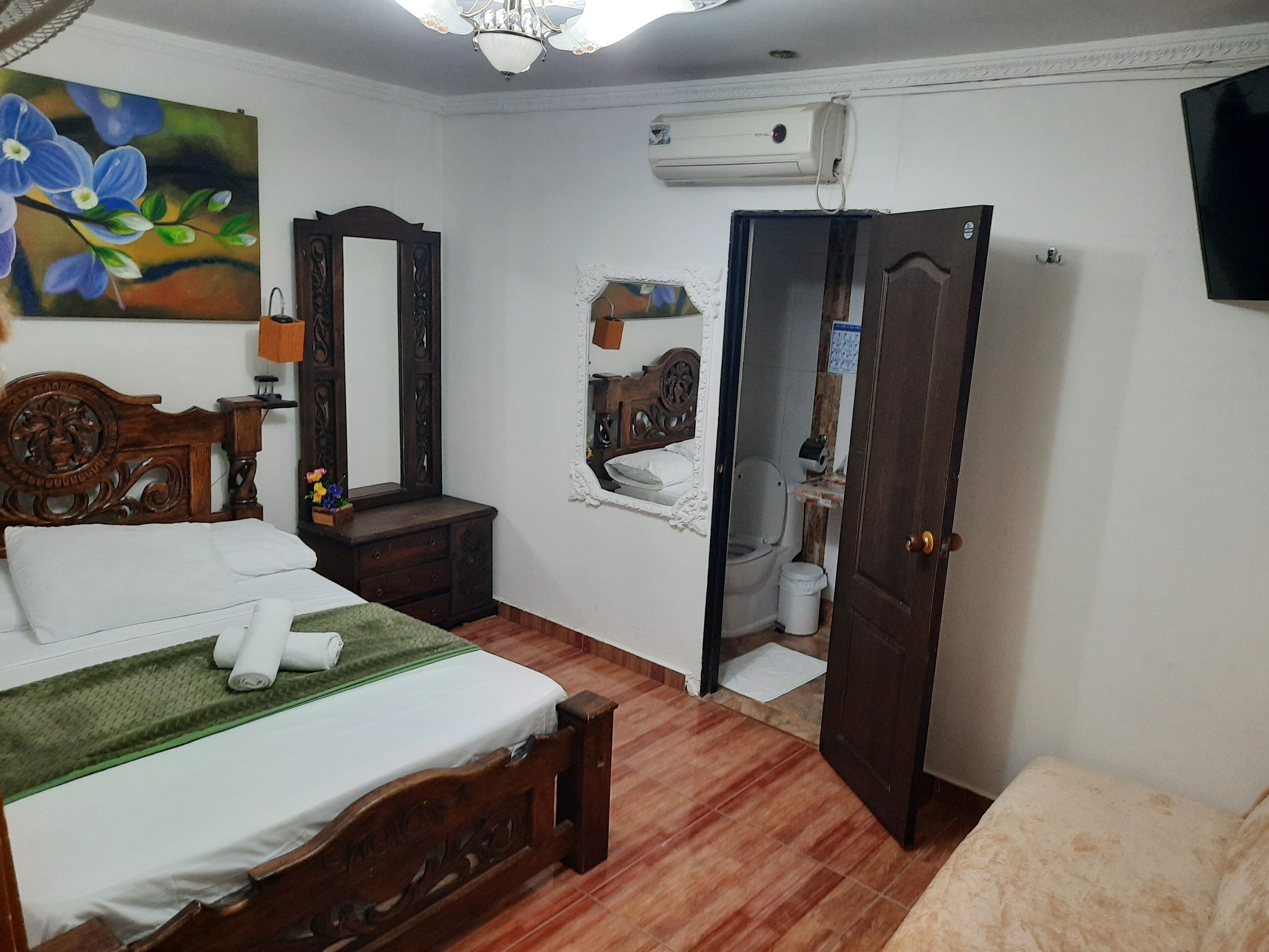 A bedroom with a wooden bed, a nightstand, a mirror, an air conditioning unit, and a door leading to a bathroom.