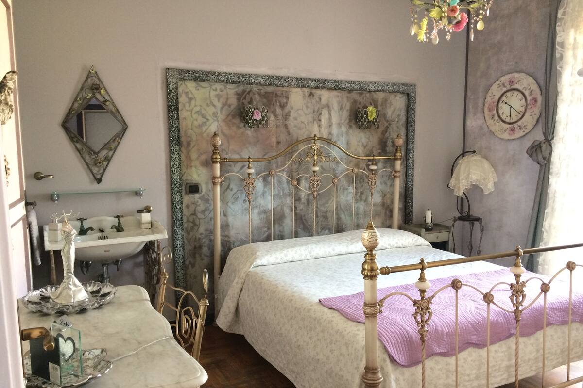 A bedroom with a vintage-style bed, a bedside table with a mirror, a dresser, and a wall clock.
