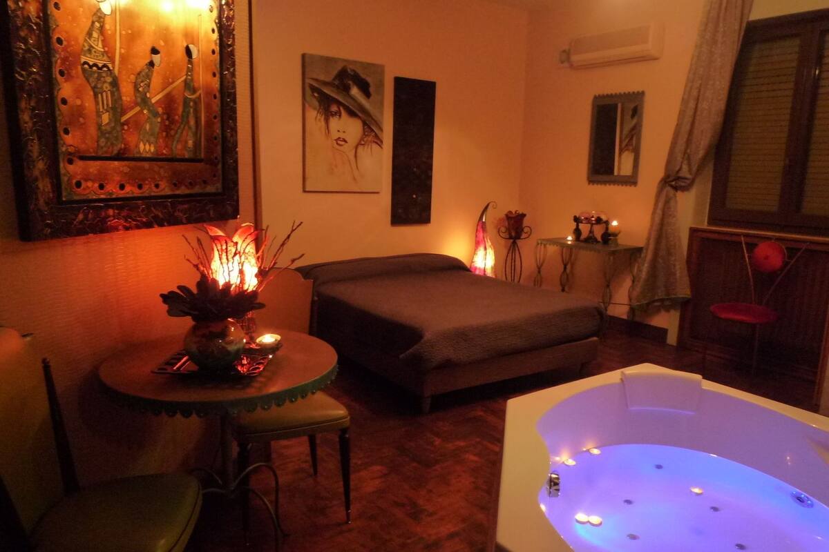 A room with a large bed, a sitting area with a table and chairs, a jacuzzi, and artwork on the walls.