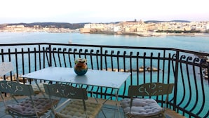 Un balcón con una mesa y dos sillas, con vistas a una masa de agua y un paisaje urbano al fondo.