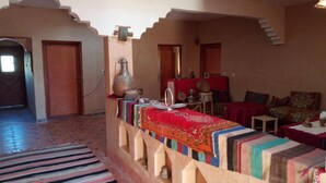 A traditional room with a long, striped rug, a sofa with a red cover, and a table with a colorful cloth. - Dar Zara (Ighrem N'Ougdal)