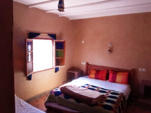 A bedroom with a bed, a window with a view, a wall-mounted shelf, and a bedside table.