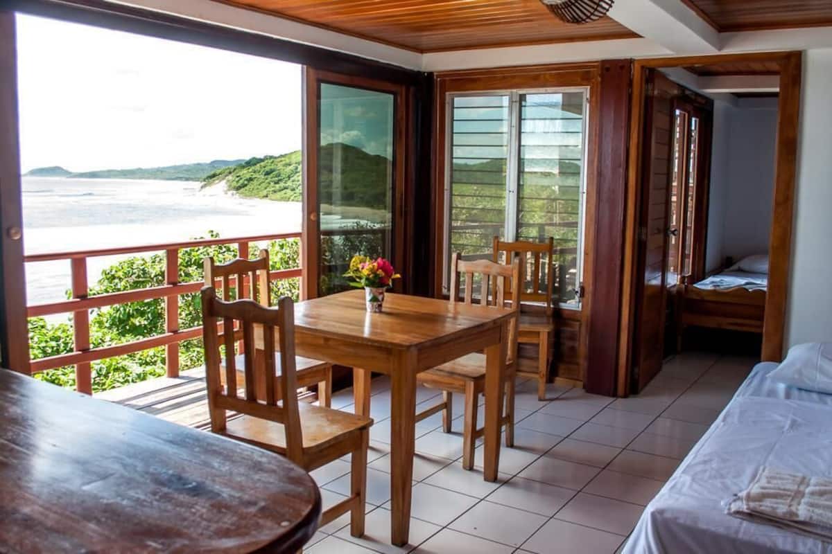 A room with a wooden table and chairs, a bed, and a view of the ocean.