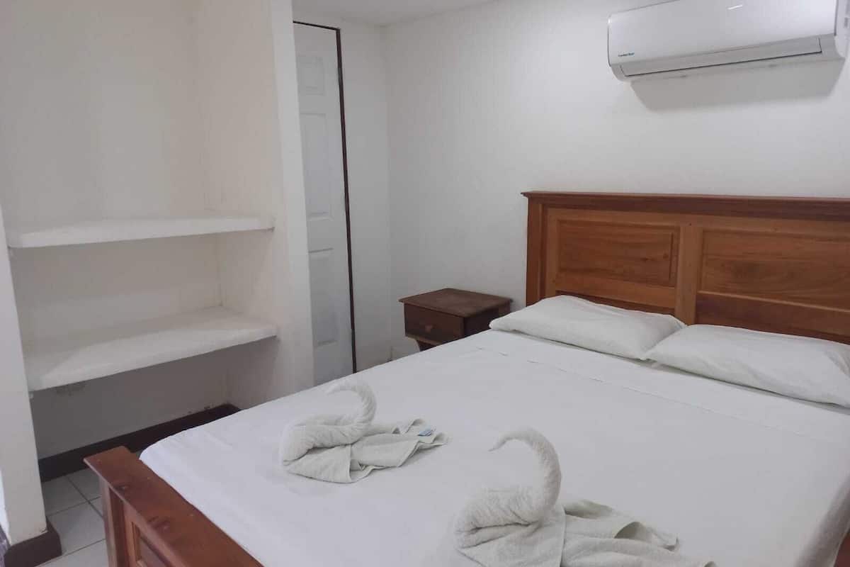 A hotel room with a wooden bed, white linens, a nightstand, a wall-mounted air conditioner, and a door.