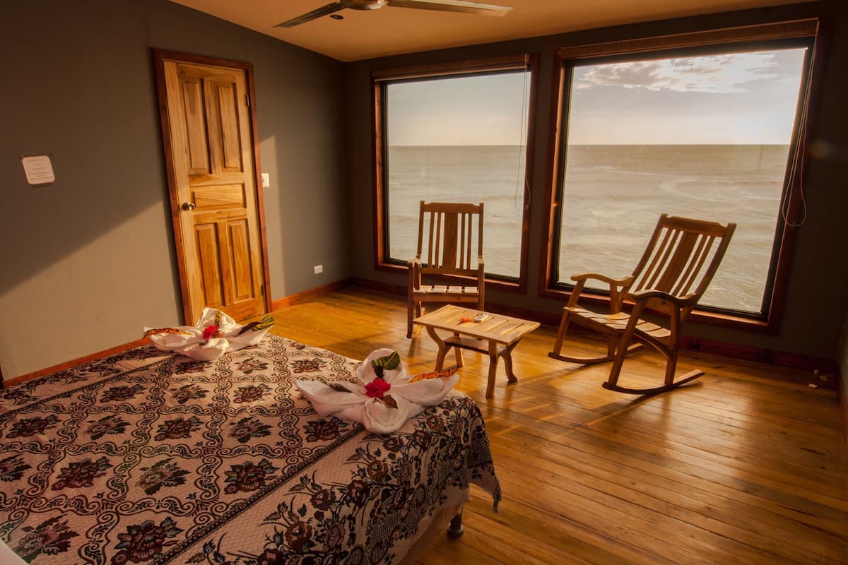A room with a bed, two rocking chairs, a small table, and a large window overlooking the ocean.