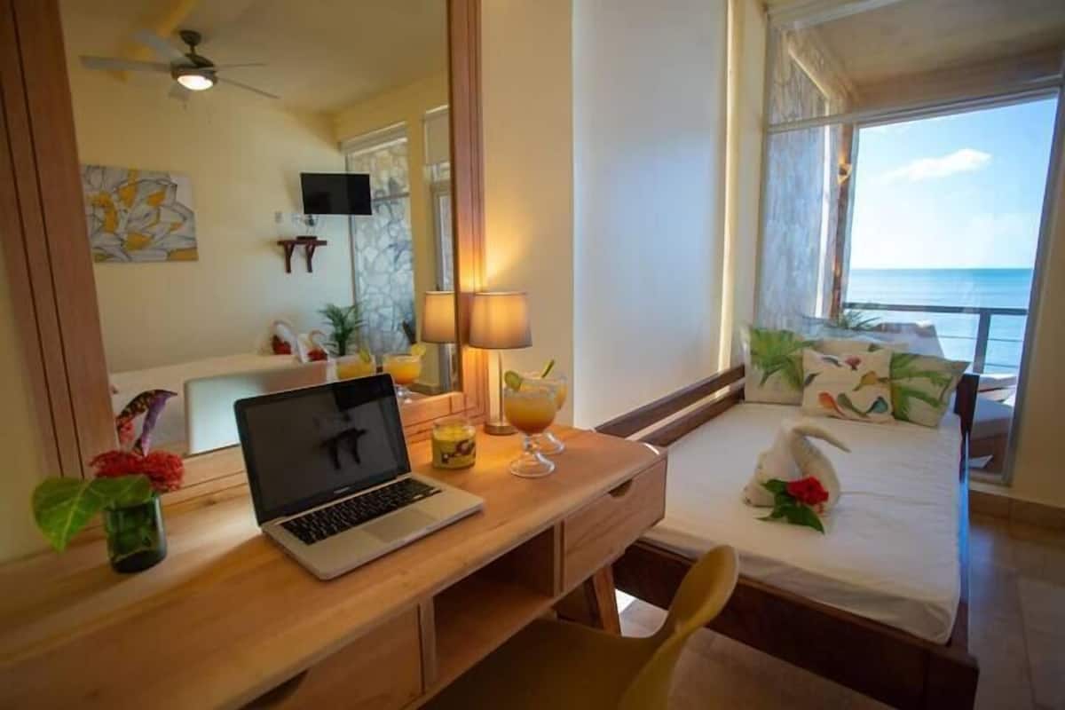 A hotel room with a wooden desk, laptop, two drinks, a swan decoration on the bed, a TV, and a view of the ocean.