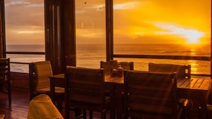 A restaurant with wooden furniture and large windows overlooking the ocean at sunset. - Magnific Rock - Surf Resort & Yoga Retreat Nicaragua - Hostel (Tola)