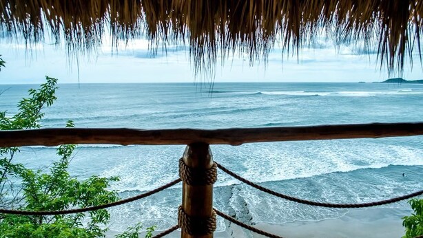 A beach view with a wooden railing and rope barrier, overlooking the ocean and a distant island. - Magnific Rock - Surf Resort & Yoga Retreat Nicaragua - Hostel (Tola)