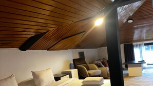 Una habitación amplia con techo alto, una cama con ropa blanca, una silla de masaje y un escritorio pequeño.