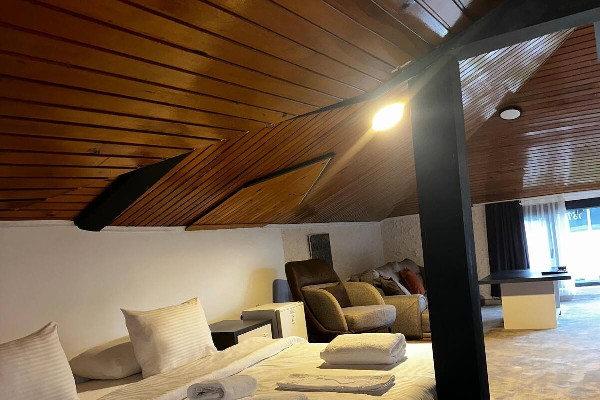 Une pièce spacieuse avec un plafond haut, un lit recouvert de draps blancs, un fauteuil de massage et un petit bureau.