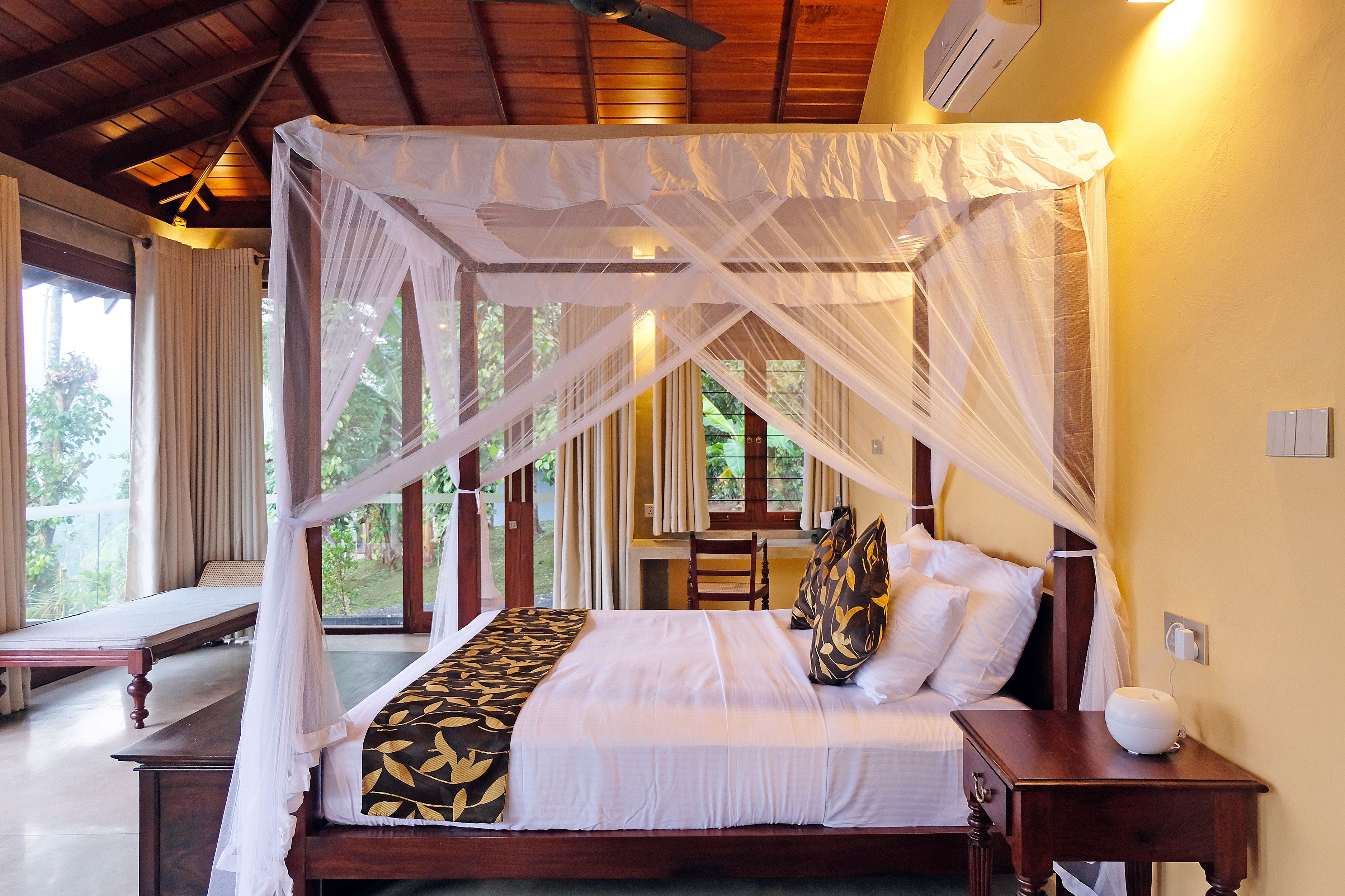 A four-poster canopy bed with a white and black bedspread, a bedside table with a white lamp, and a view of the outdoors through large windows.