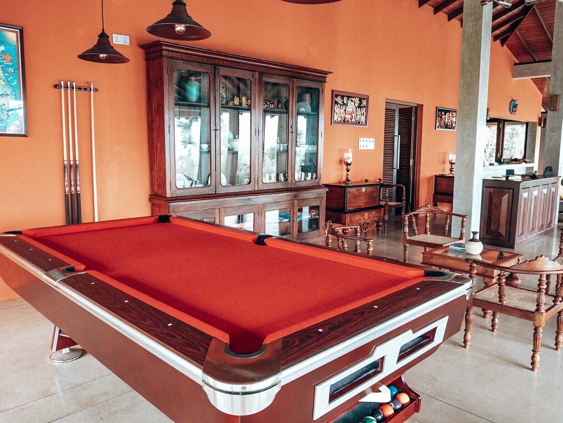 A pool table with a red felt top, wooden cabinets, and a dining area with chairs.