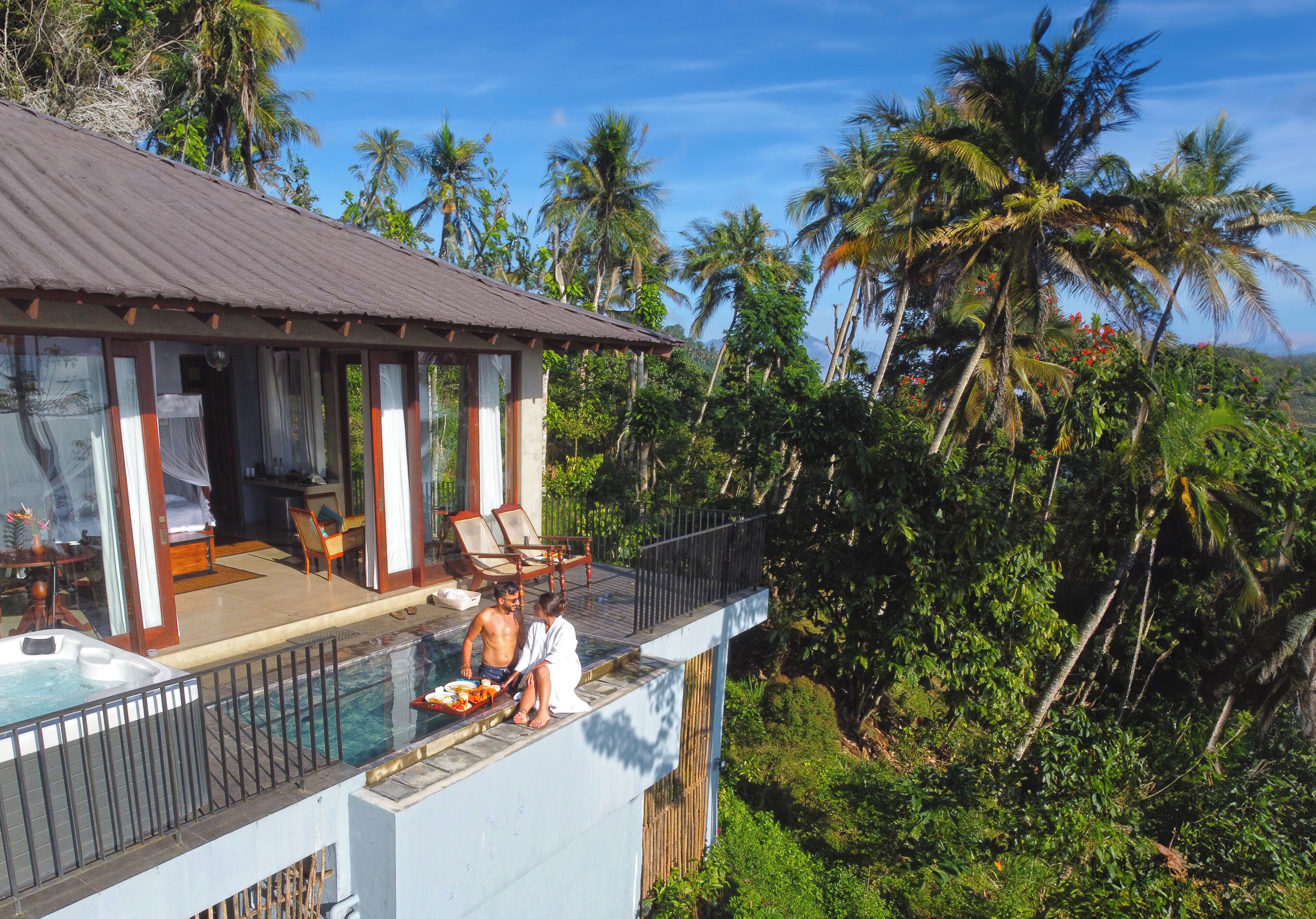 A two-story building with a swimming pool on the upper level, surrounded by lush greenery and palm trees.